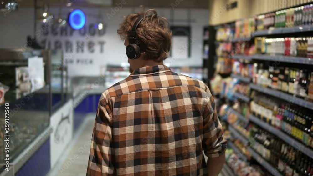 Stylish caucasian guy with headphones walks through goods section of the store, wearing plaid shirt - listening to the good music.. Following back view shot. Slow Motion Stylish caucasian guy with headphones walks through goods section of the store, wearing plaid shirt - listening to the good music.. Following back view shot. Slow Motion