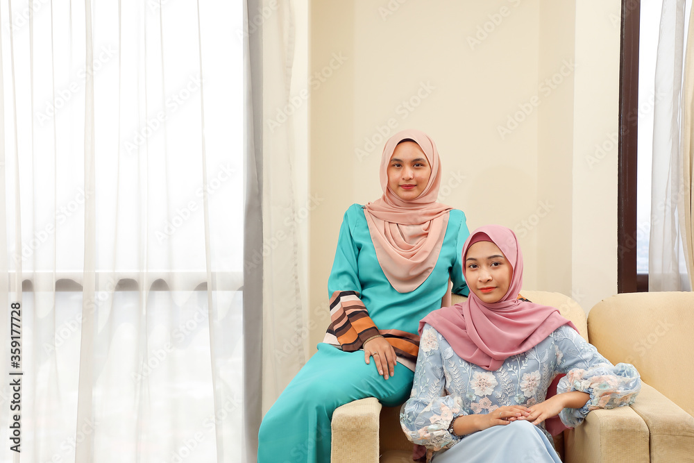Two young Asian Malay Muslim woman wearing headscarf at home office student sitting on sofa look at camera Two young Asian Malay Muslim woman wearing headscarf at home office student sitting on sofa look at camera