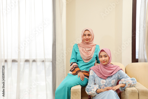 Two young Asian Malay Muslim woman wearing headscarf at home office student sitting on sofa look at camera