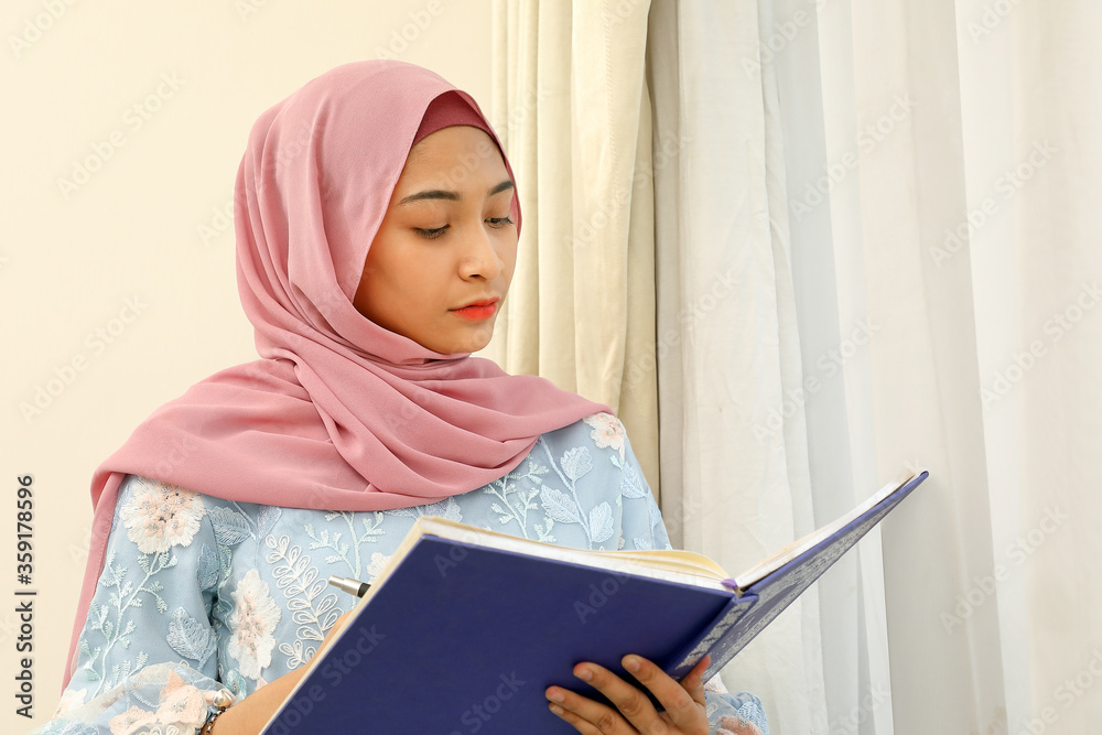 young Asian Malay Muslim woman wearing headscarf at home office next to window look at book document study think write young Asian Malay Muslim woman wearing headscarf at home office next to window look at book document study think write