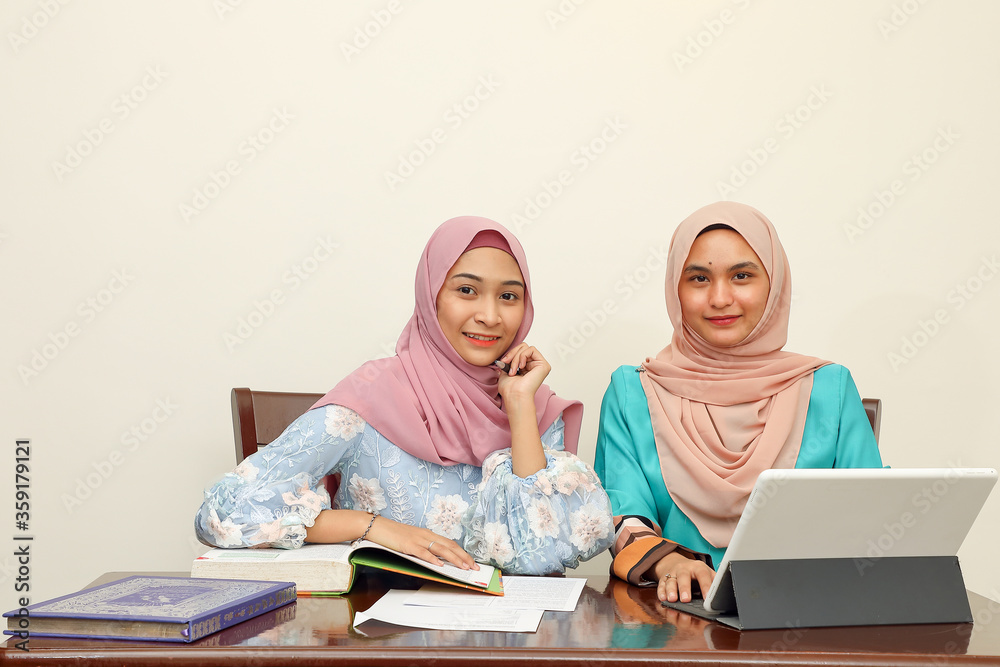Two young Asian Malay Muslim woman wearing headscarf at home office student sitting at table talk mingle look at computer book document study discuss smile happy Two young Asian Malay Muslim woman wearing headscarf at home office student sitting at table talk mingle look at computer book document study discuss smile happy