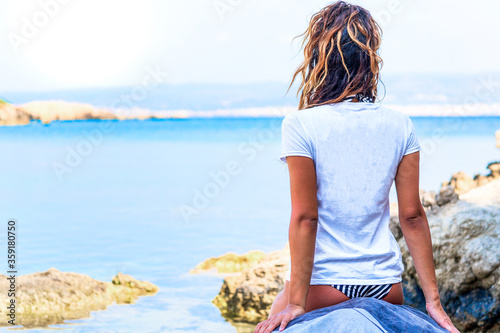 A girl with blonde hair in the foreground of shoulders with a white T-shirt observes the sea.