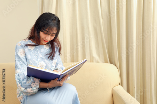 young Asian Malay Muslim woman wearing baju kurung dress at home sit rest on sofa read book notes write with pen think © oqba