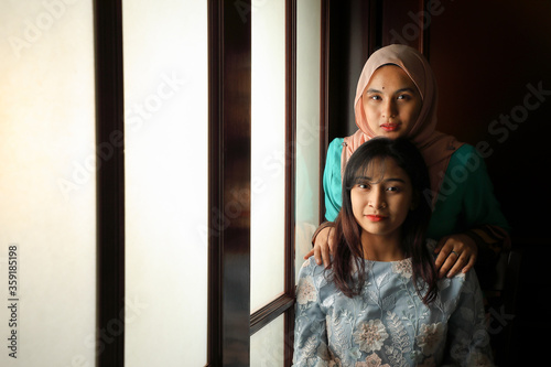two young Asian Malay muslim woman wearing headscarf baju kurung dress at home in front window mood light on face dark background peaceful content calm tranquil break trough wonder happy © oqba