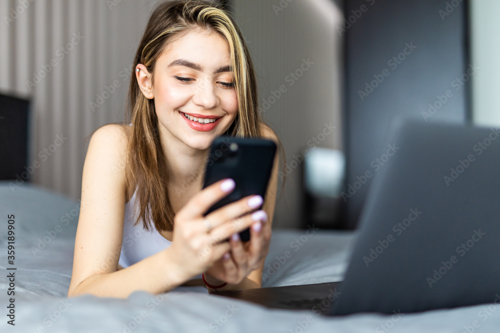 Beautiful smiling woman lying on the bed by the laptop and typing a message on her phone. Beautiful smiling woman lying on the bed by the laptop and typing a message on her phone.