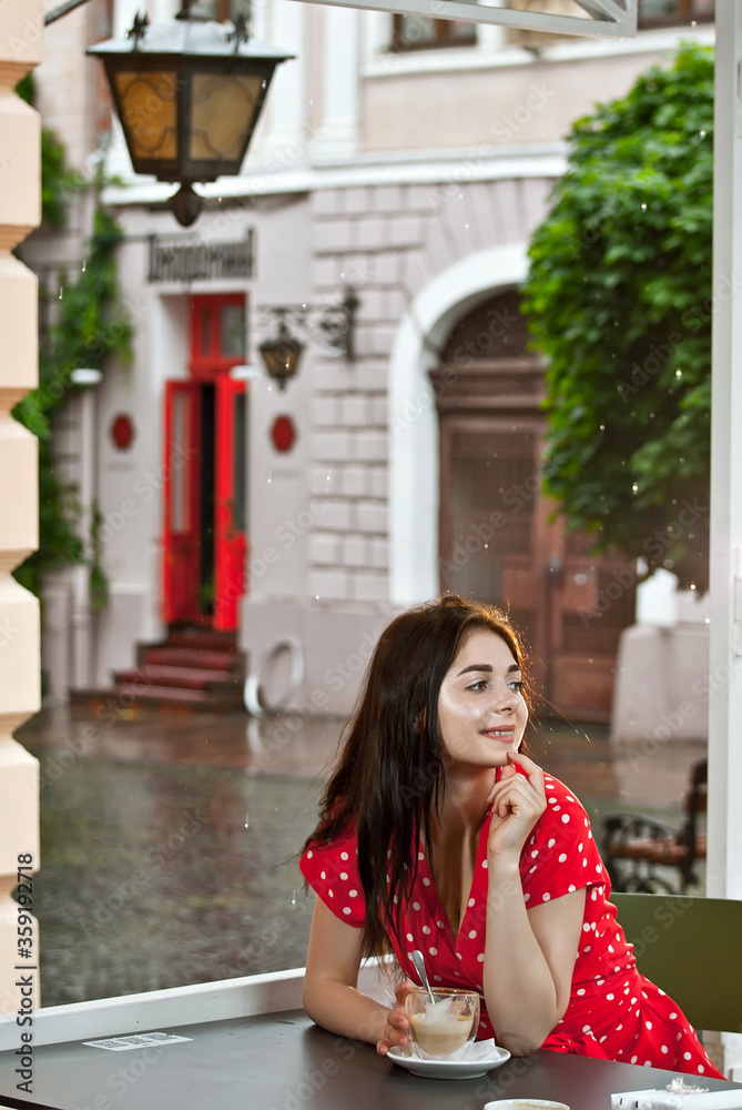 Happy girl sitting at the table of a street cafe. Woman drinks coffee in a red in a white dot dress. Happy girl sitting at the table of a street cafe. Woman drinks coffee in a red in a white dot dress.