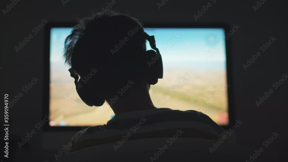Boy plays game of War Thunder strategy online on computer with headphones in the evening in room. Light of monitor and silhouette of child play emotionally at computer Boy plays game of War Thunder strategy online on computer with headphones in the evening in room. Light of monitor and silhouette of child play emotionally at computer