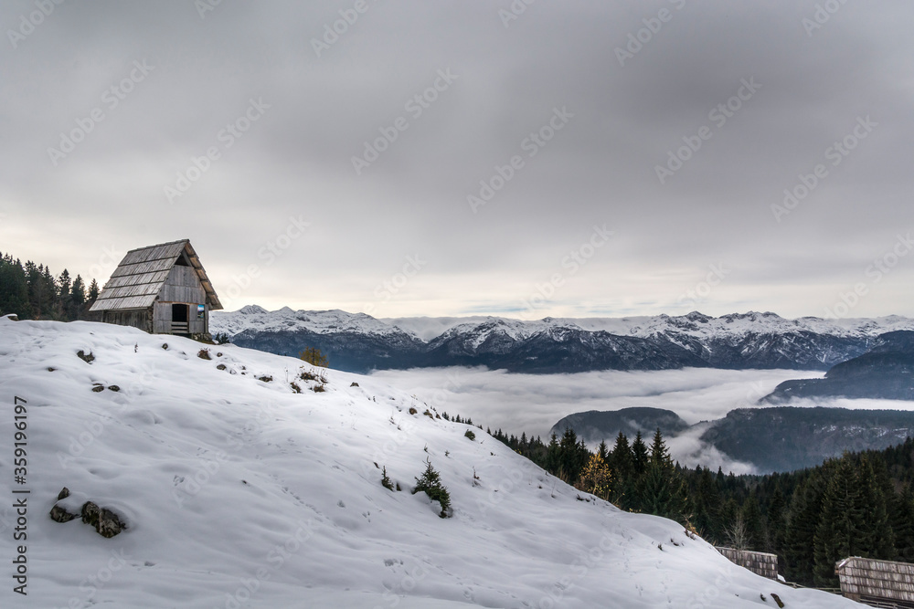 Beautiful snowy landscape in Slovenia. Zajamniki village in winter season. Alps mountains covered in snow. Small wooden hut on top. Inversion clouds fog in valley and cloudy sky