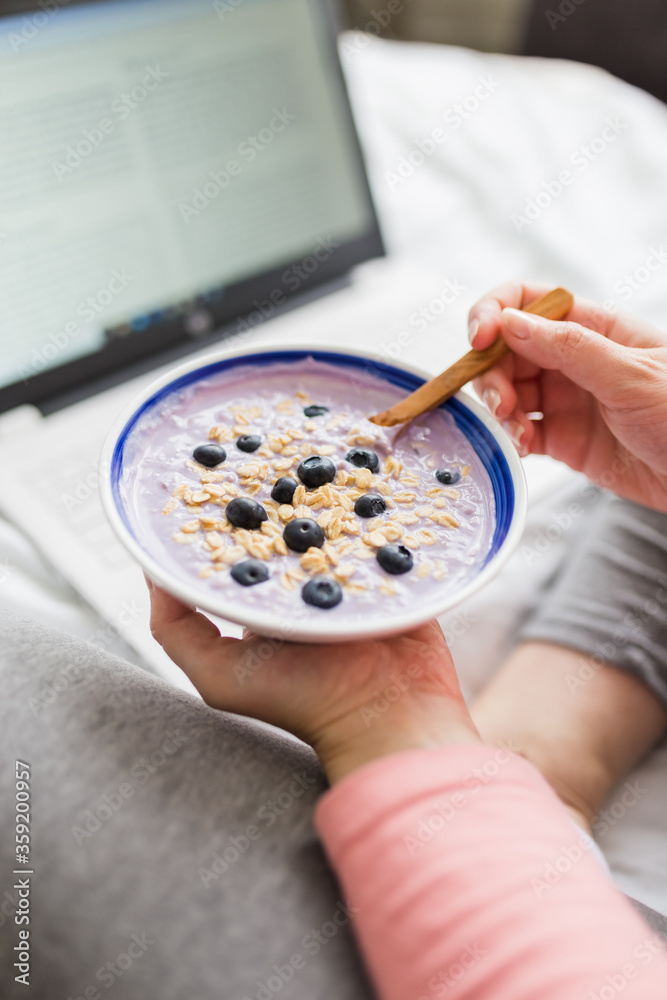 Close up of pregnant woman eating muesli with blueberries while working on laptop.