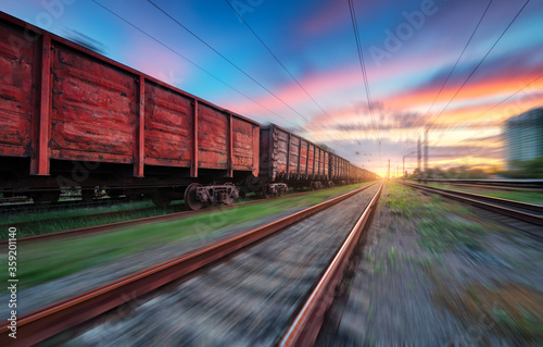 Moving freight train at sunset. Railroad and beautiful sky with clouds with motion blur effect in summer. Industrial landscape with train, railway station and blurred background. Railway platform