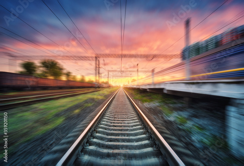 Railroad and beautiful sky with clouds at sunset with motion blur effect in summer. Industrial landscape with freight train, railway station and blurred background. Railway platform in speed motion