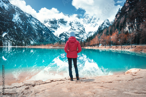 Young woman in red jacket is standing on shore of lake with azure water in autumn in Dolomites, Italy. Landscape with girl, reflection in water, snowy mountains, blue sky with clouds, trees. Travel