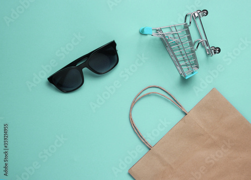 Minimalism shopping concept. Paper shopping bag, mini shopping cart and sunglasses on blue background. Top view