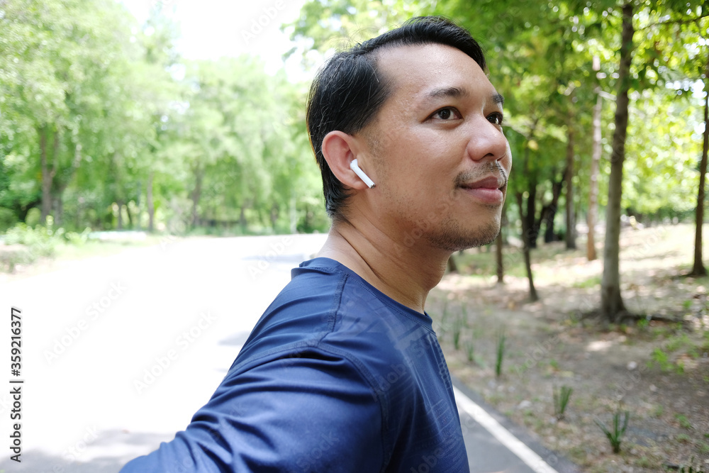 Smiling Asian sportman in sportwear is running or jogging and listen to music with white headphones in the garden. Smiling Asian sportman in sportwear is running or jogging and listen to music with white headphones in the garden.