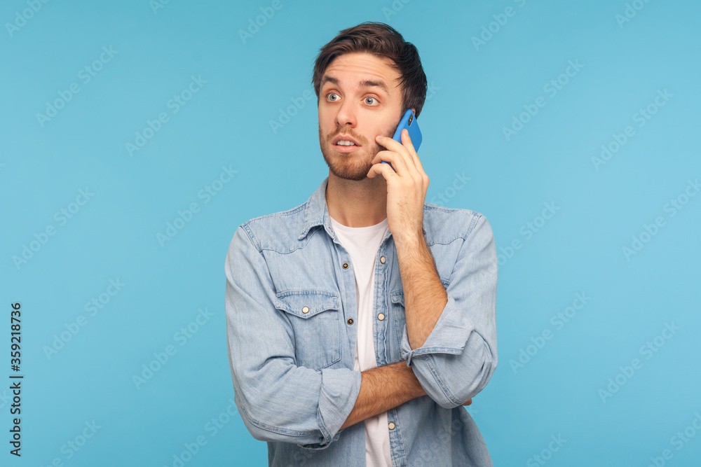 Portrait of amazed young businessman in casual denim shirt talking on phone and looking shocked, surprised by unexpected mobile conversation with partner. studio shot isolated on blue background Portrait of amazed young businessman in casual denim shirt talking on phone and looking shocked, surprised by unexpected mobile conversation with partner. studio shot isolated on blue background