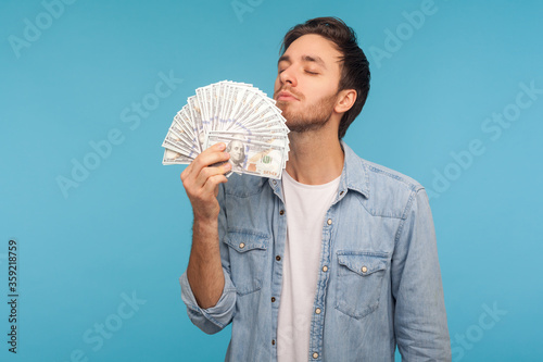 Portrait of greedy man in worker denim shirt smelling dollar banknotes with pleasure expression, enjoying big money profit and rich life, lottery win. indoor studio shot isolated on blue background