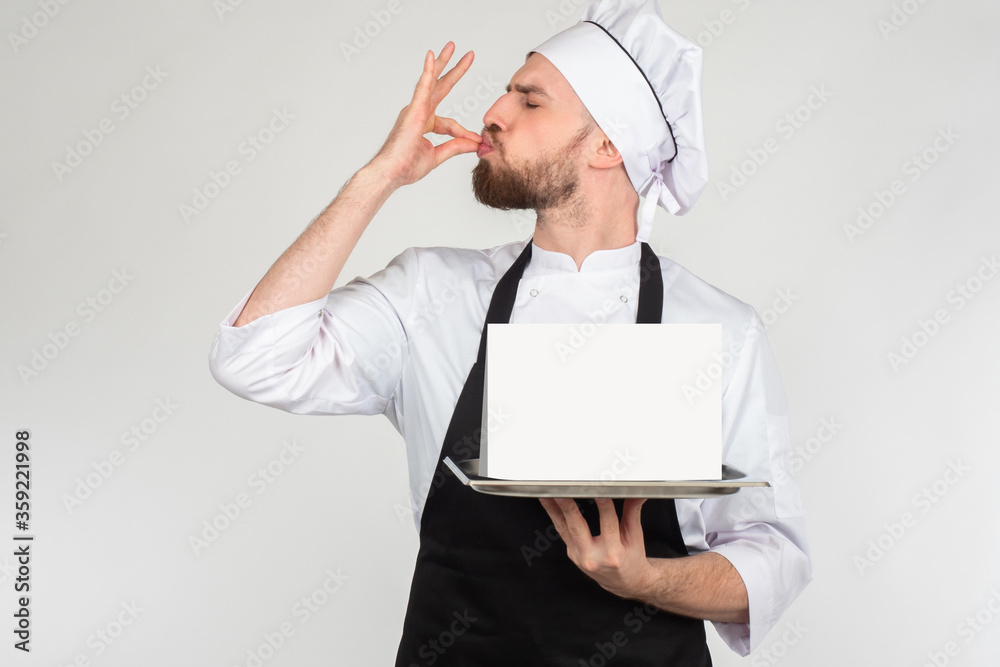 The gesture means delicious. The chef holds a tray with an empty sign. Space for text. The chef makes an approving gesture with his hand. The taste of the product is highly appreciated. The gesture means delicious. The chef holds a tray with an empty sign. Space for text. The chef makes an approving gesture with his hand. The taste of the product is highly appreciated.