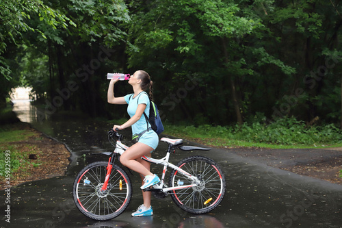 sport girl in sportswear stands with her back beside a bicycle drinks water from a bottle