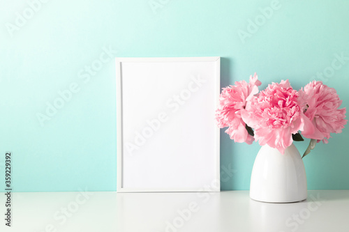 Home decoration with floral decor, frame poster on table. Beautiful flowers pink peonies in vase on mint background