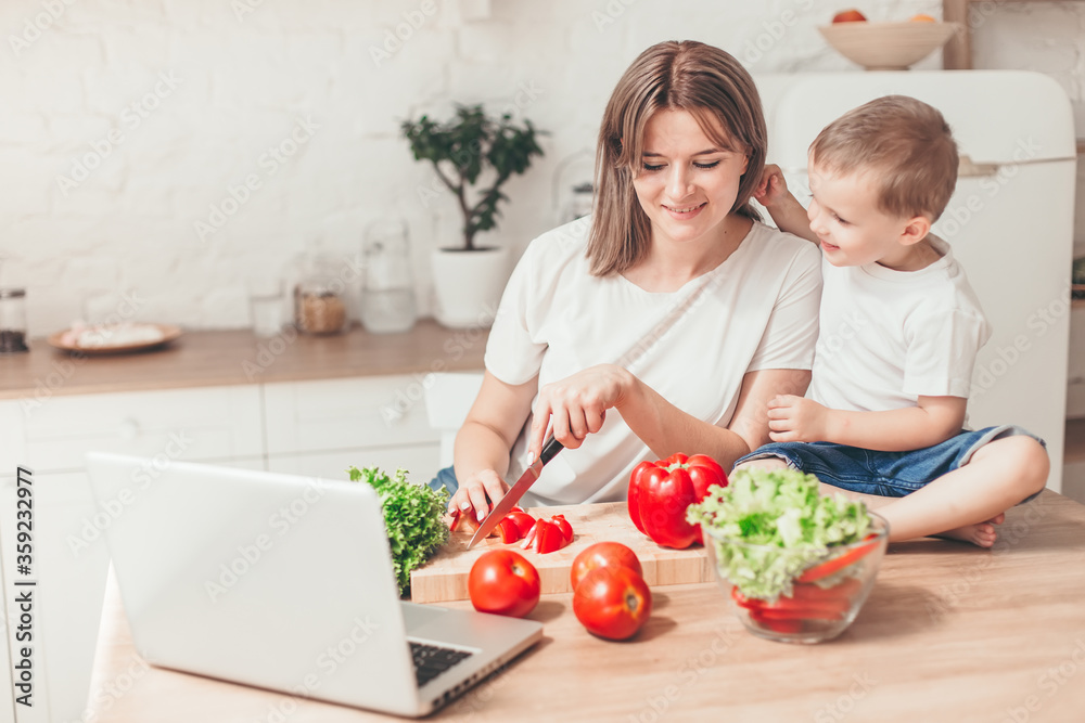 Son with mom are cutting vegetables on salad in kitchen and have a good time together. Happy family.