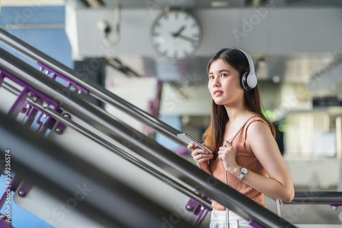 Obraz na płótnie Young Asian woman passenger wearing headphone and using smart mobile phone to listening music and walking up the stairs in subway station when traveling in big city, commuter and transportation concep w salonie