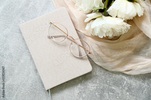 Flat lay composition with paper notebook, glasses, bouquet of peonies flowers and beige fabric on concrete table. Top view stylish feminine workspace. photo