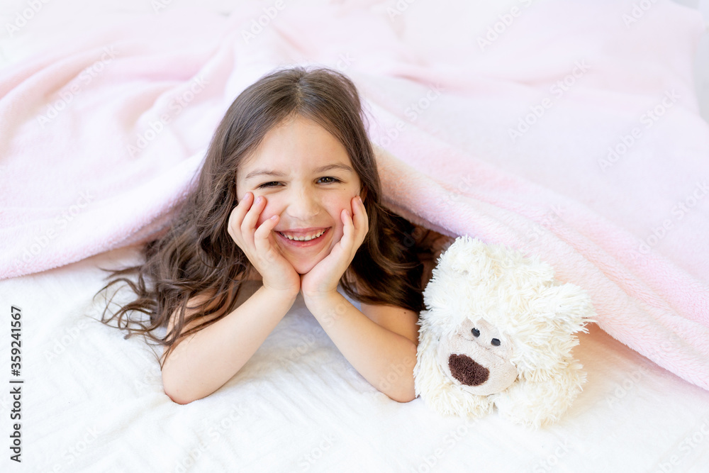 a small smiling girl 5-6 years old is lying in bed with a Teddy bear, hands under her cheeks