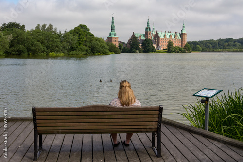 Obraz na płótnie Blond girl sitting on a bench and looking at a beautiful Frederiksborg Castle and a lake around it, in Hillerød, Denmark w salonie
