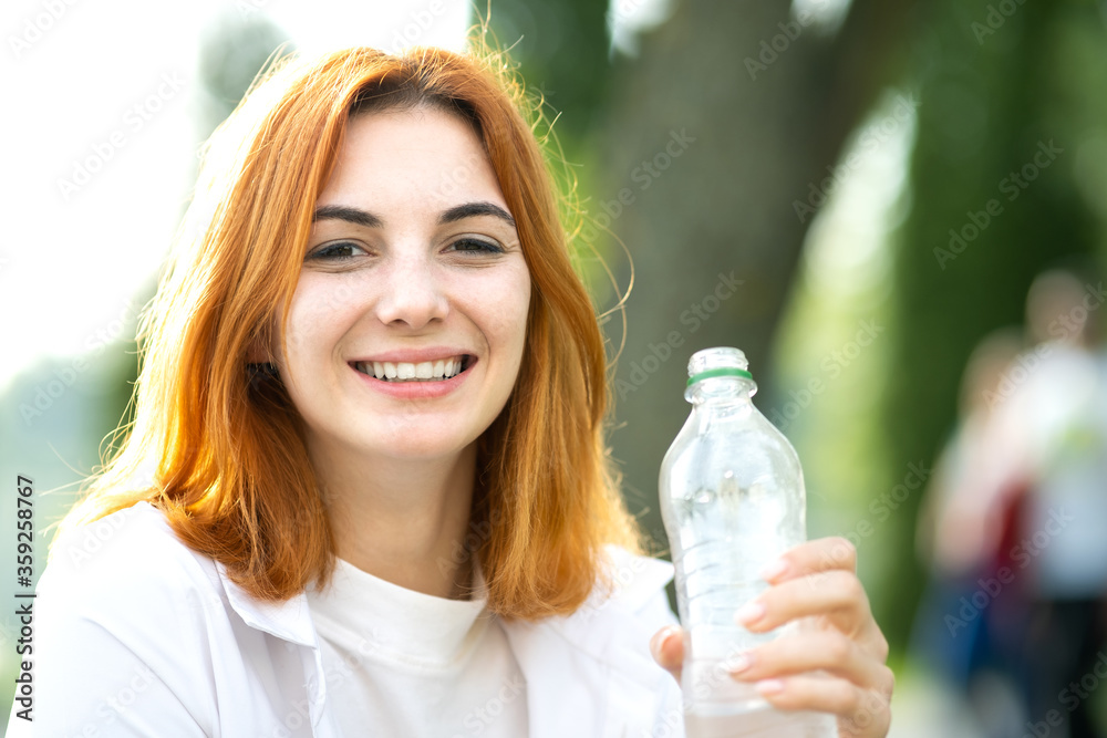 Thirsty smiling redhead woman drinking water from a bottle in summer park. Thirsty smiling redhead woman drinking water from a bottle in summer park.