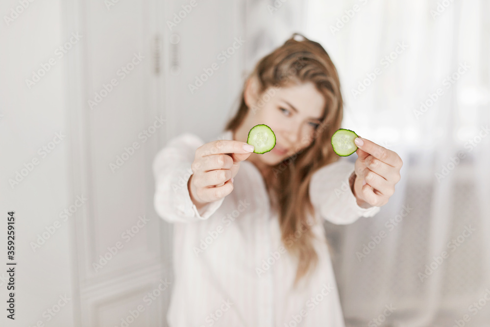 A cheerful girl holds cucumber slices in her hands for a face mask. Skin care. The concept of beauty and health. Blurred background A cheerful girl holds cucumber slices in her hands for a face mask. Skin care. The concept of beauty and health. Blurred background
