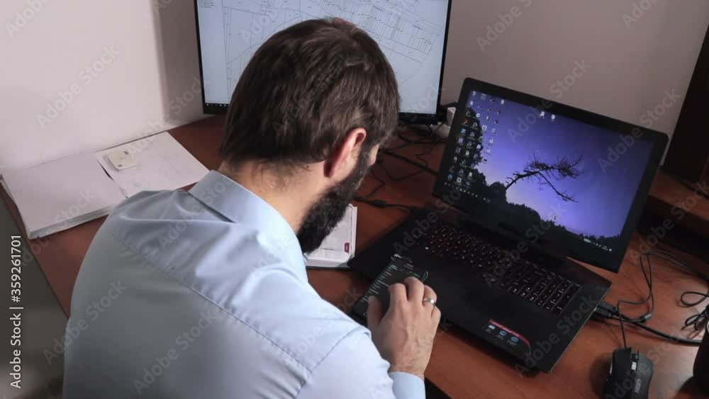 Young man sitting at his desk checking his phone working in front of the laptop in the office slider shot to the right from above Young man sitting at his desk checking his phone working in front of the laptop in the office slider shot to the right from above