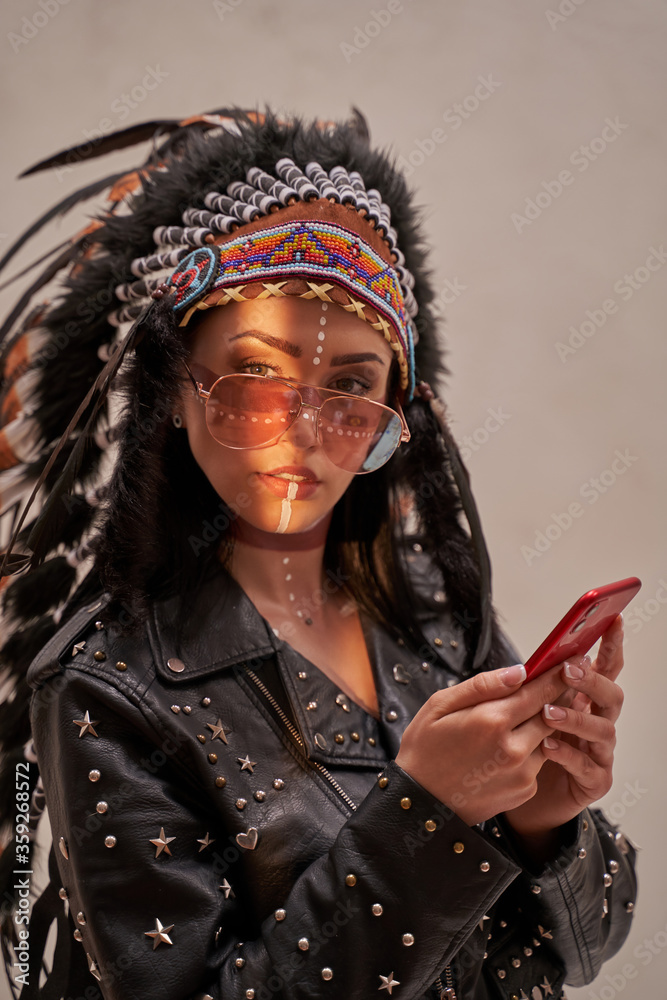 Beautiful and confident female wearing American indian traditional headdress, leather jacket and sunglasses while posing in a bright studio and looking serious while typing a message on her phone