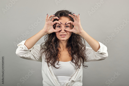 Making glasses of fingers, looking at camera and smiling, funny, hold hands near eyes, imitating binoculars, holding arm eyebrows, through imaginary binocular, grimacing, mask with hand. Young woman