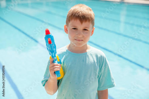 Cute little blond boy in blue t-short with toy water gun staying near the swimming pool. Having fun on vacation playing water gun fight.