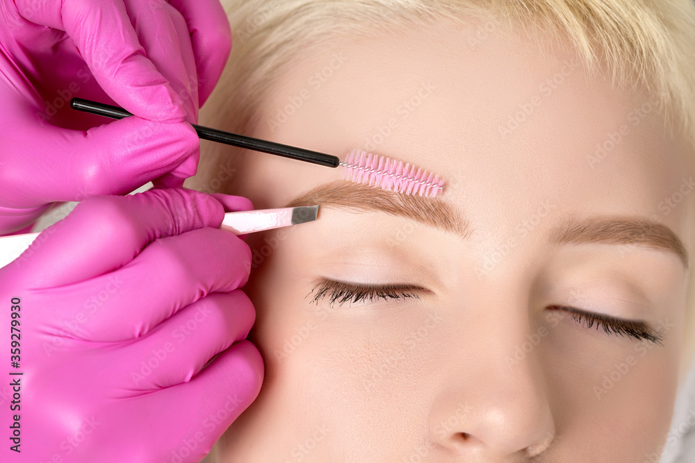 Makeup artist plucking eyebrows with tweezers to a beautiful woman with short blonde hair and with healthy clean skin. Women's cosmetology in the beauty salon.