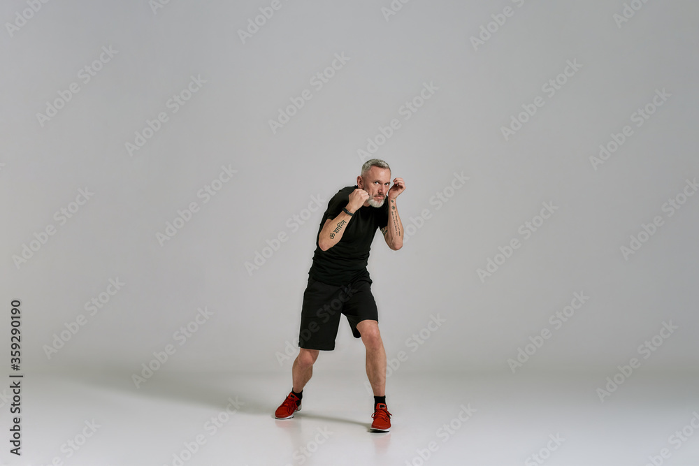 Full length shot of middle aged muscular man in black sportswear practicing punches while exercising in studio over grey background