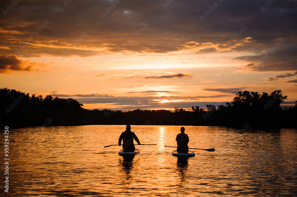beautiful rear view of two sitting people on sup boards which floating on the river at sunset