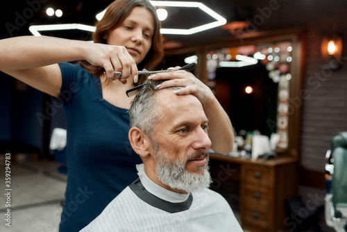 Modern male hairstyle. Handsome bearded mature man sitting in armchair in the barbershop while young barber girl doing haircut for him. Man visiting beauty salon