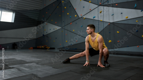 Full length shot of active young man in sportswear looking aside, while stretching body against climbing wall. Concept of sport life and rock climbing