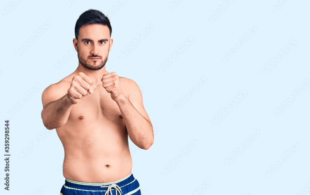 Fototapeta premium Young handsome man wearing swimwear punching fist to fight, aggressive and angry attack, threat and violence