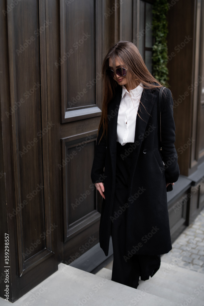 Outdoor photo of brunette lady walking on street background in cold autumn day. Fashion street style portrait. wearing dark casual trousers, white shirt, sunglasses and black coat. Fashion concept. Outdoor photo of brunette lady walking on street background in cold autumn day. Fashion street style portrait. wearing dark casual trousers, white shirt, sunglasses and black coat. Fashion concept.