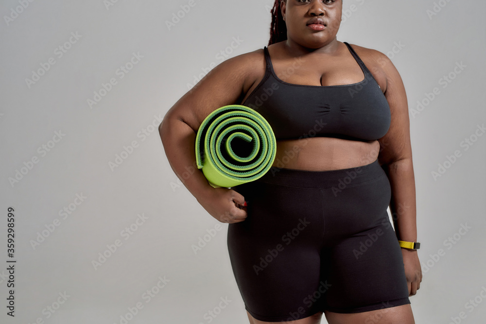 Yoga time. Cropped shot of plump, plus size african american woman in  sportswear holding green yoga mat for fitness, posing in studio over grey  background Stock Photo