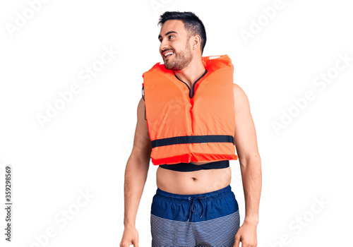 Young handsome man wearing lifejacket looking away to side with smile on face, natural expression. laughing confident.