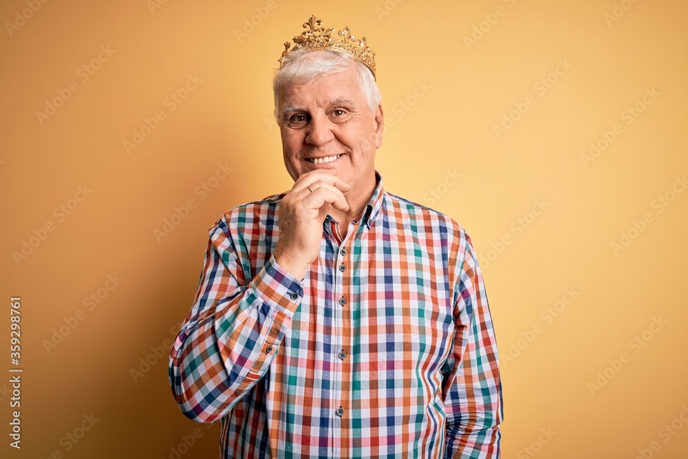 Senior handsome hoary man wearing golden crown of king over isolated yellow background looking confident at the camera with smile with crossed arms and hand raised on chin. Thinking positive. Senior handsome hoary man wearing golden crown of king over isolated yellow background looking confident at the camera with smile with crossed arms and hand raised on chin. Thinking positive.