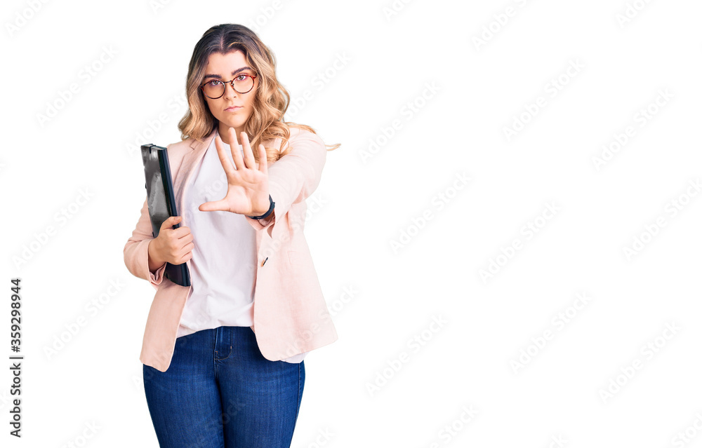 Young caucasian woman wearing business clothes and glasses holding binder with open hand doing stop sign with serious and confident expression, defense gesture Young caucasian woman wearing business clothes and glasses holding binder with open hand doing stop sign with serious and confident expression, defense gesture