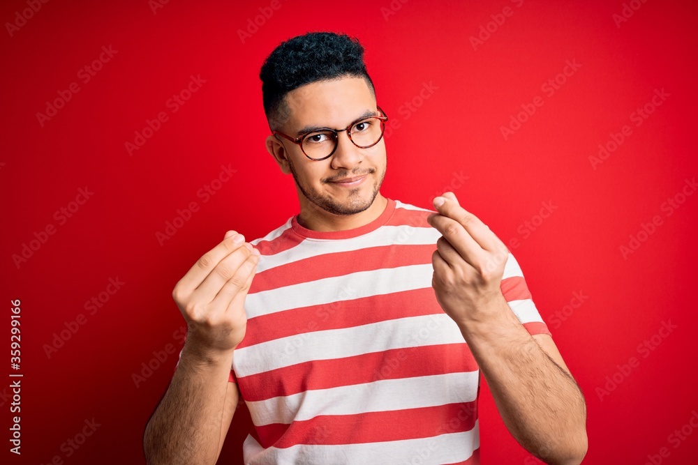 Young handsome man wearing casual striped t-shirt and glasses over isolated red background doing money gesture with hands, asking for salary payment, millionaire business Young handsome man wearing casual striped t-shirt and glasses over isolated red background doing money gesture with hands, asking for salary payment, millionaire business