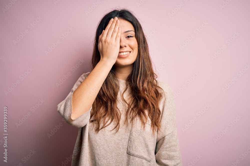 Young beautiful brunette woman wearing casual sweater standing over pink background covering one eye with hand, confident smile on face and surprise emotion. Young beautiful brunette woman wearing casual sweater standing over pink background covering one eye with hand, confident smile on face and surprise emotion.