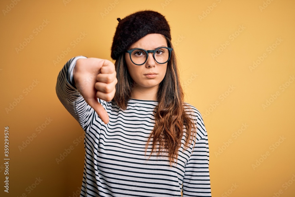 Young beautiful brunette woman wearing french beret and glasses over yellow background looking unhappy and angry showing rejection and negative with thumbs down gesture. Bad expression. Young beautiful brunette woman wearing french beret and glasses over yellow background looking unhappy and angry showing rejection and negative with thumbs down gesture. Bad expression.