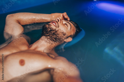 Handsome beard man floating in tank filled with dense salt water used in meditation, therapy, and alternative medicine.