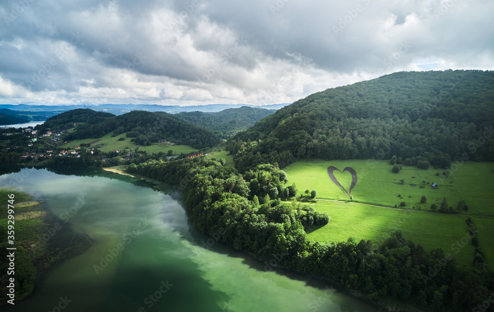 Fototapeta premium Heart shape on green hill surrounded by mountains, trees, river and lonely empty road. Love sign in memory of a lost loved one in Myczkowce, Bieszczady Mountains, Poland.
